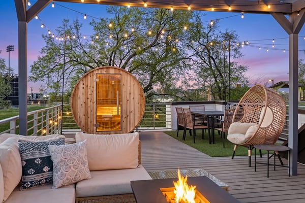 Rooftop deck at dusk with wooden sauna, fire pit, hanging lights, lounge seating, and large oak tree overhead.